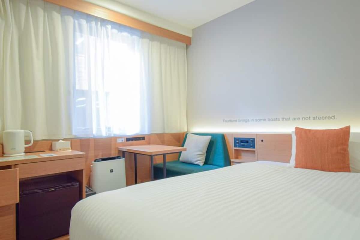A hotel room with a bed, a desk, a chair, a small table, a trash bin, and a window with curtains.