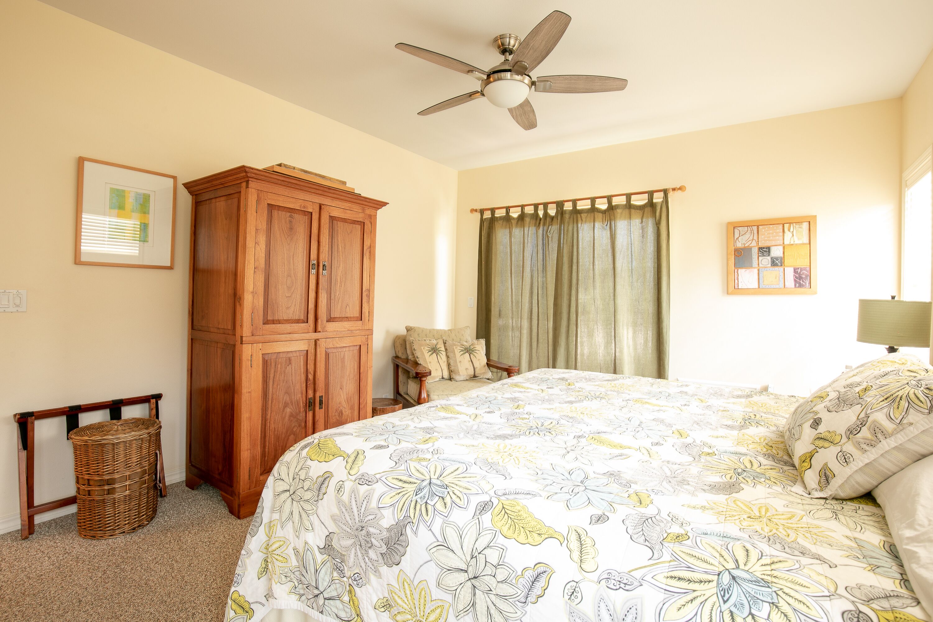 Un dormitorio con cama, ventilador de techo, armario de madera, cesta de mimbre y ventana con cortinas.