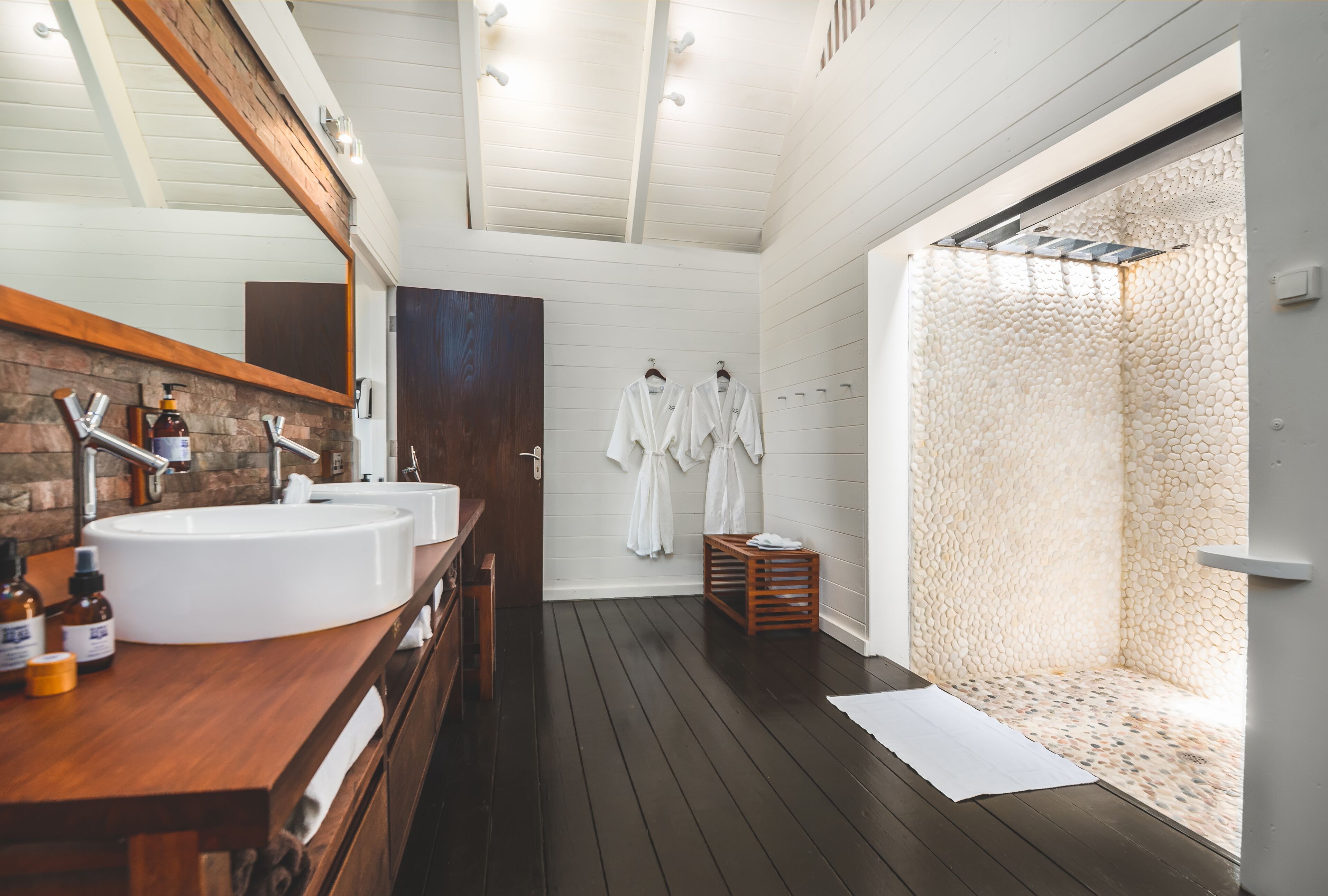 A modern bathroom with two sinks, wooden vanity, and a walk-in shower with a pebble wall.