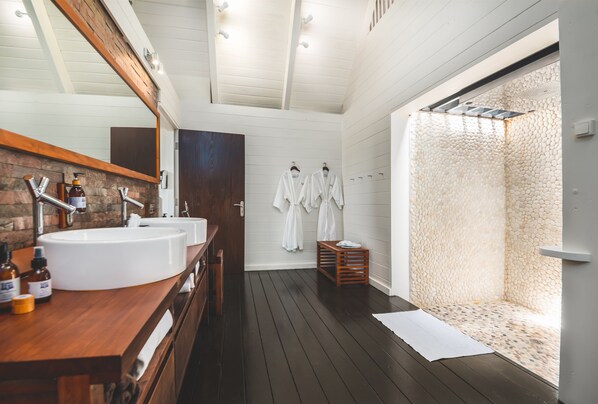 A modern bathroom with two sinks, wooden vanity, and a walk-in shower with a pebble wall.