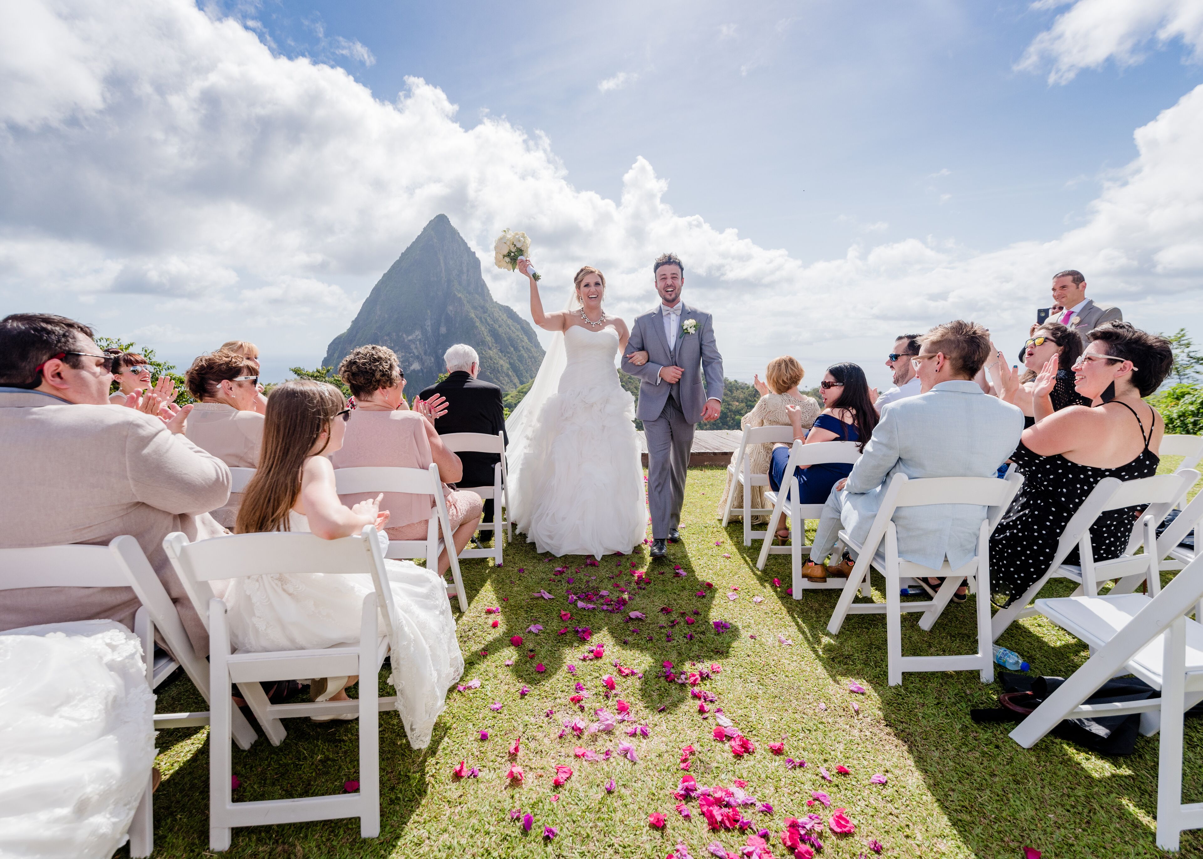 Una ceremonia de boda al aire libre, con invitados sentados en sillas blancas, una novia y un novio caminando por un pasillo, y una montaña al fondo.