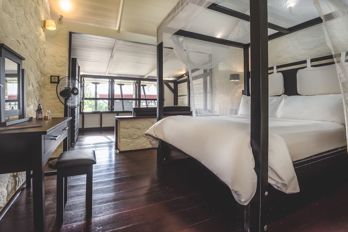 Una cama con dosel con ropa de cama blanca, un escritorio de madera con cajones, un banco y un ventanal con vistas a zonas verdes.