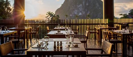 An outdoor restaurant with tables set for dining, a mountain view in the background, and a wooden fence separating the dining area from the landscape.