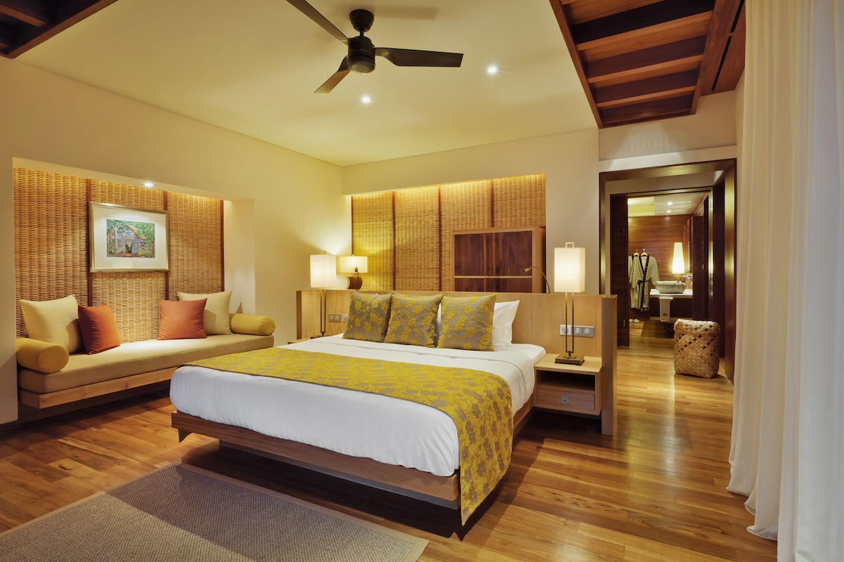 A bedroom with a bed, bedside tables, a ceiling fan, and a view into another room.