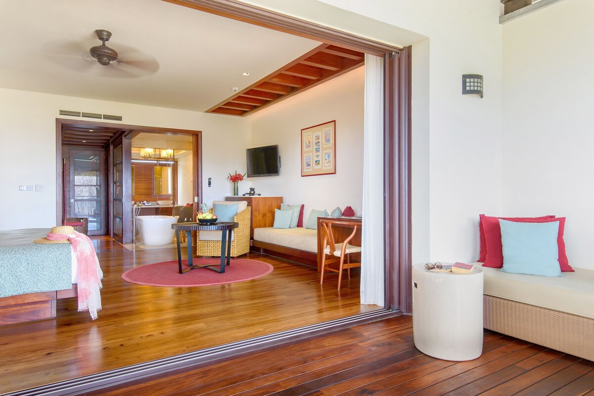 A room with a wooden floor, a sofa, a small table with a vase, a television, and a ceiling fan.