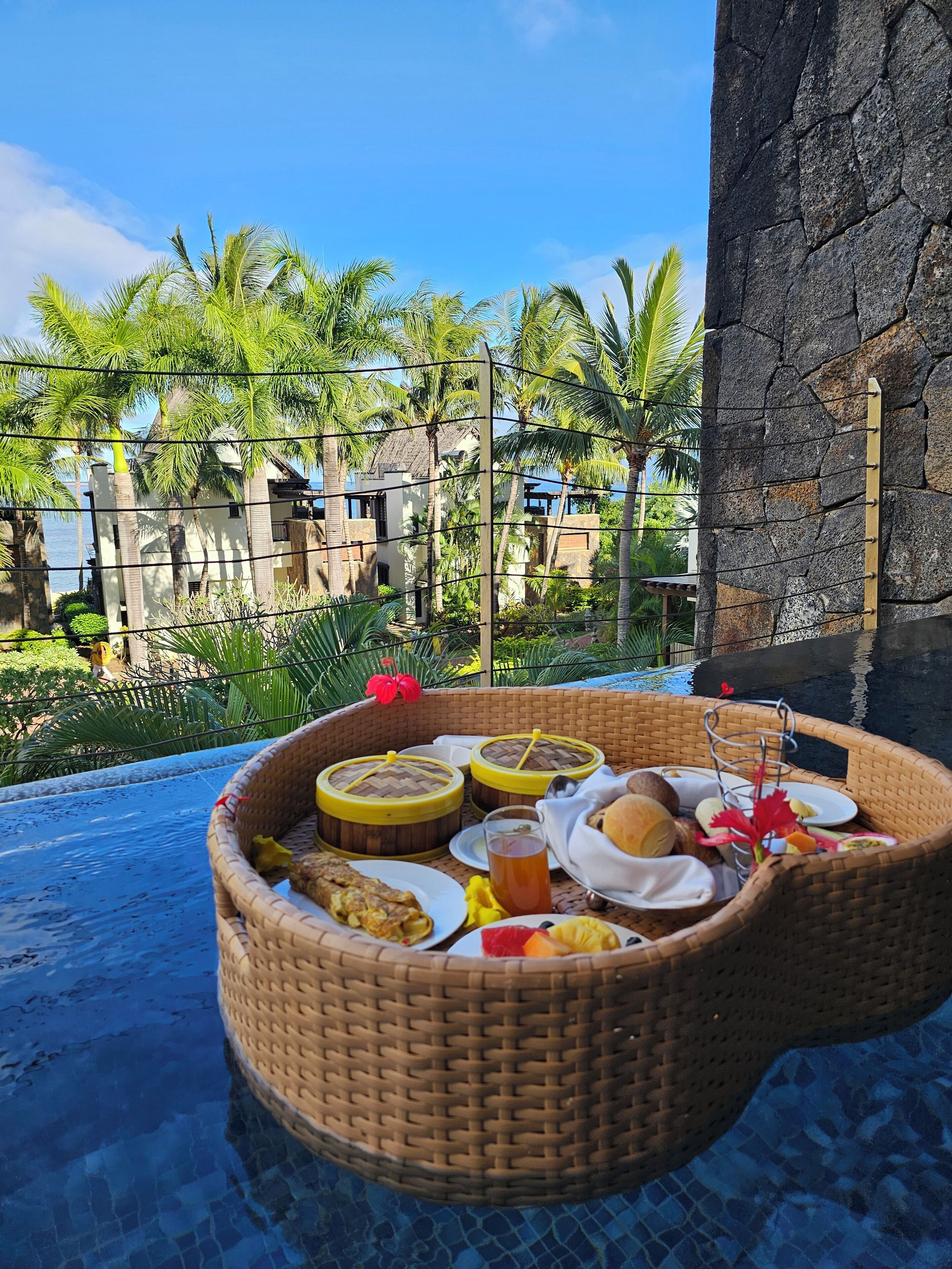 Una cesta de mimbre llena de comida y bebidas en una zona junto a la piscina, con vista a un resort y palmeras.