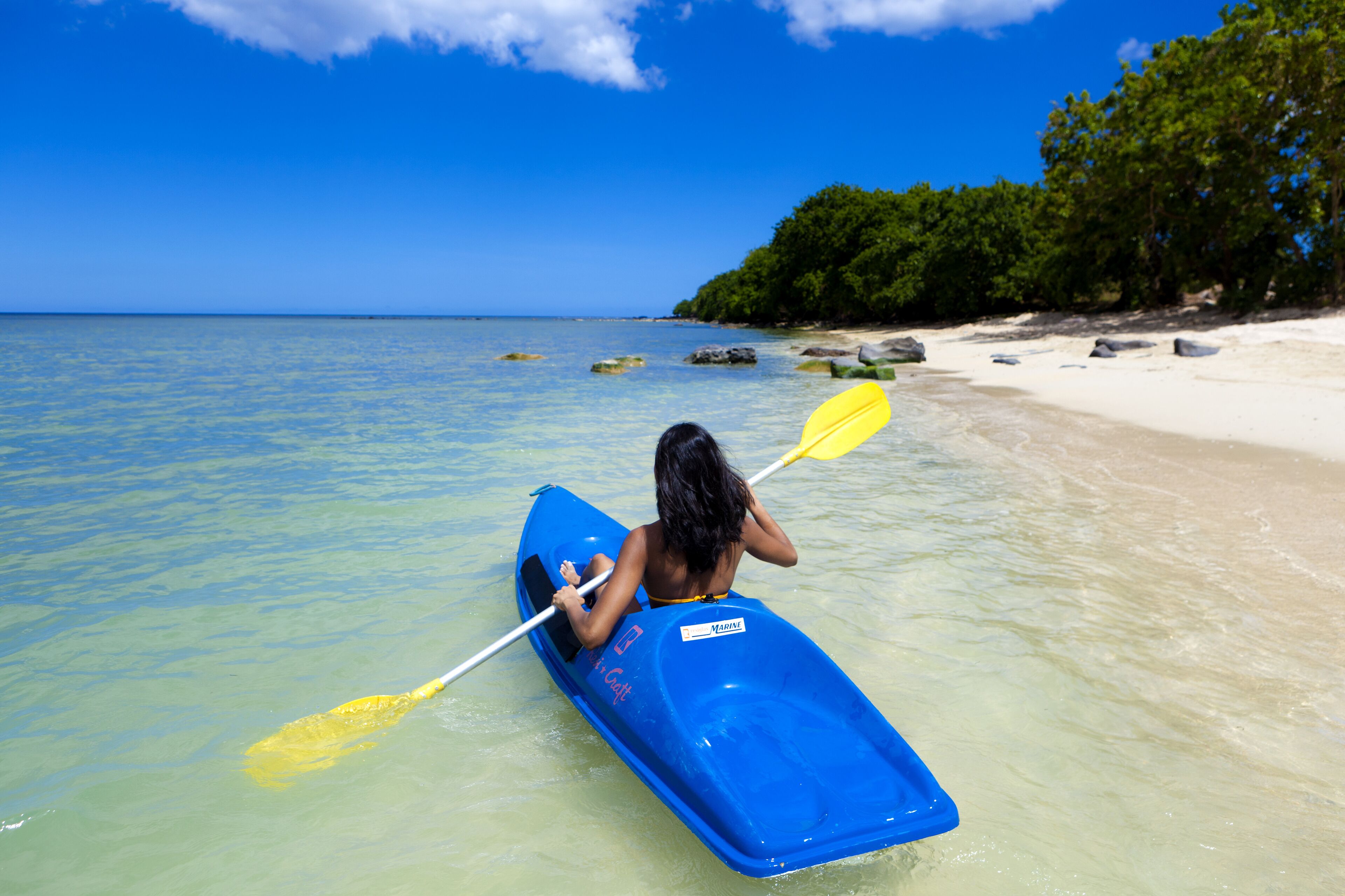 Une personne dans un kayak bleu, sur une plage où l’eau est claire et la rive bordée d’arbres.