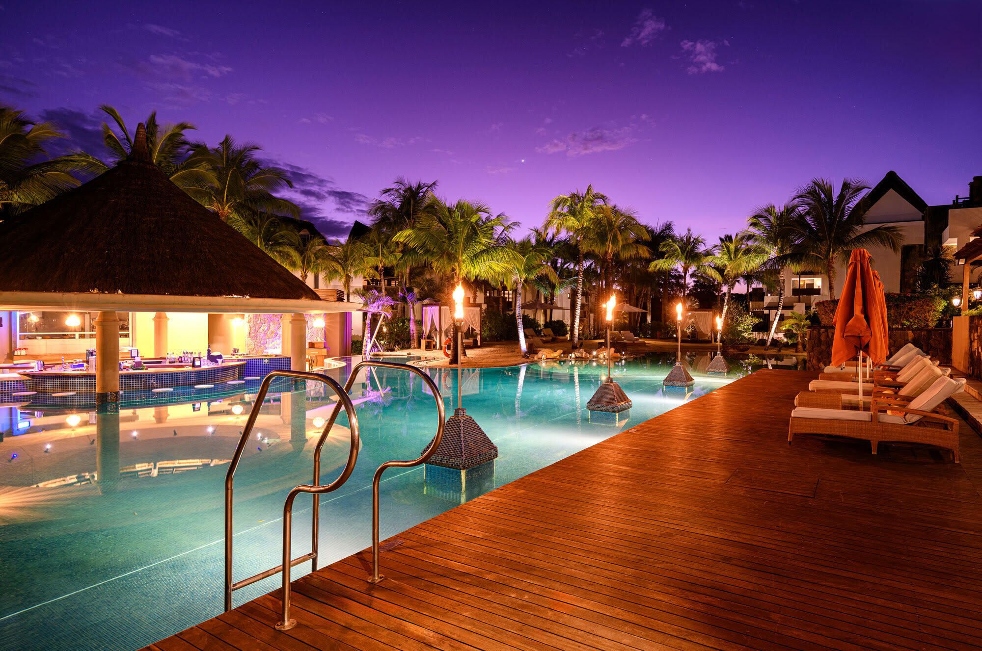 A pool area with lounge chairs and a bar, surrounded by palm trees and a thatched-roof structure.