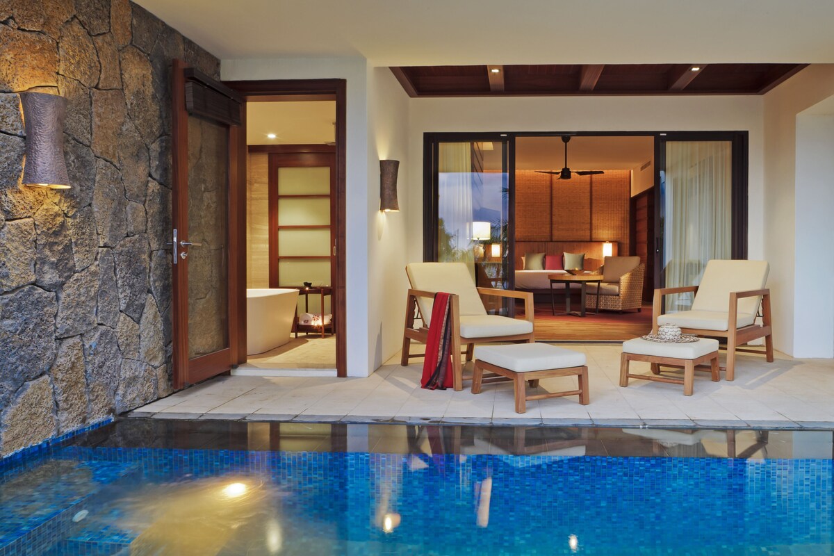 A swimming pool with lounge chairs, a stone wall, and a view into a dining area.
