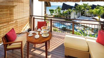 Una terraza de madera con una mesa dispuesta para tomar té, un sofá de mimbre y vistas a una piscina y edificios de un complejo turístico.