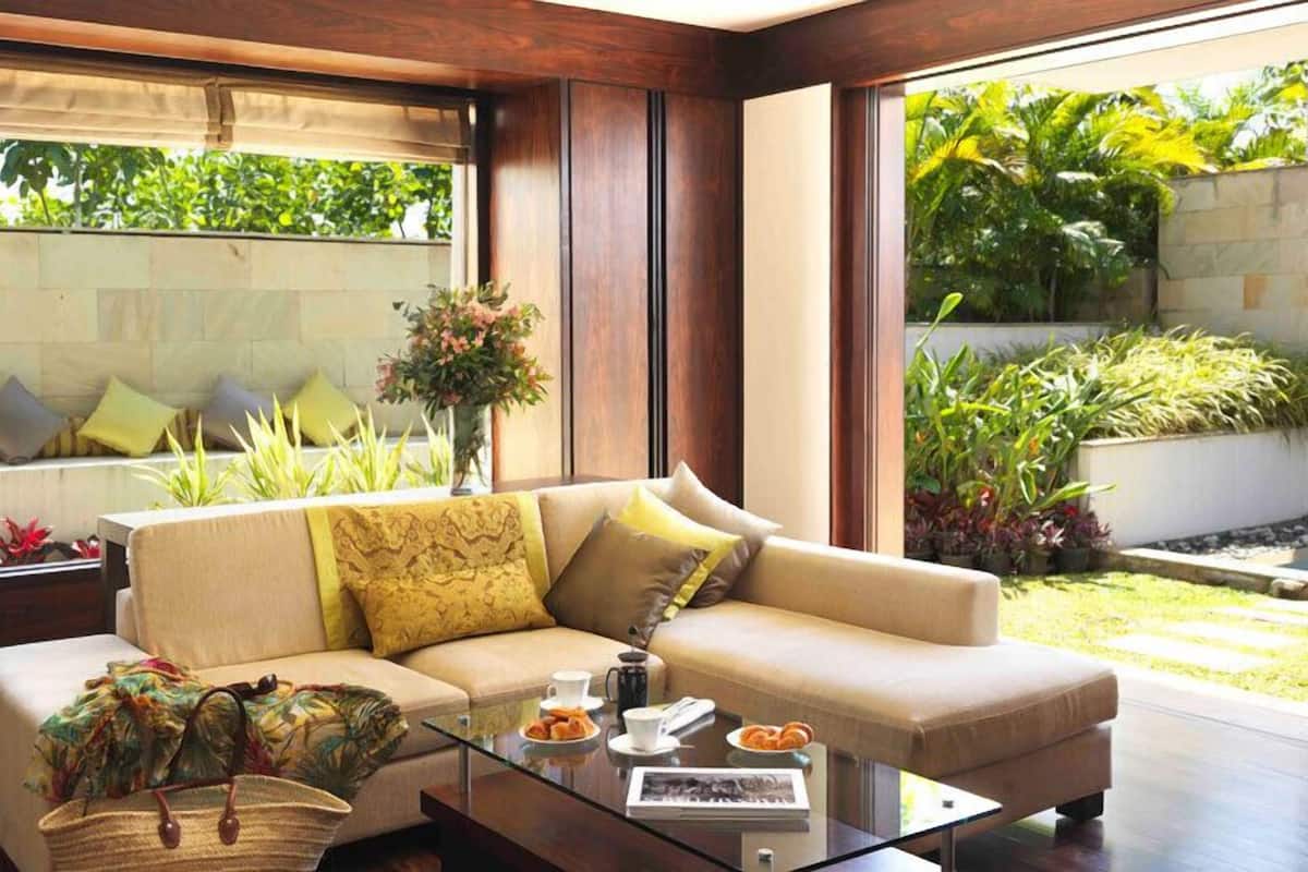 A modern living room with a beige sofa, a glass coffee table, and a view of a garden with various plants.