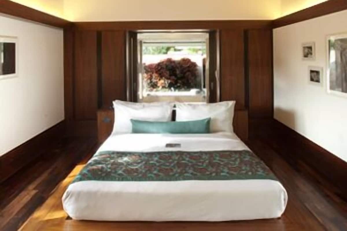 A bedroom with a wooden ceiling, a bed with white and teal bedding, and a window with a view of greenery.