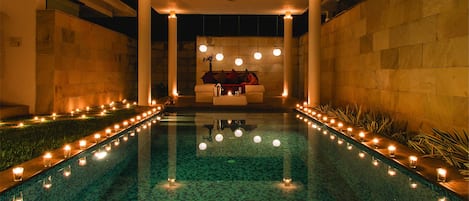 Zona de piscina con disposición de asientos bajo una estructura cubierta, rodeada de velas y plantas.