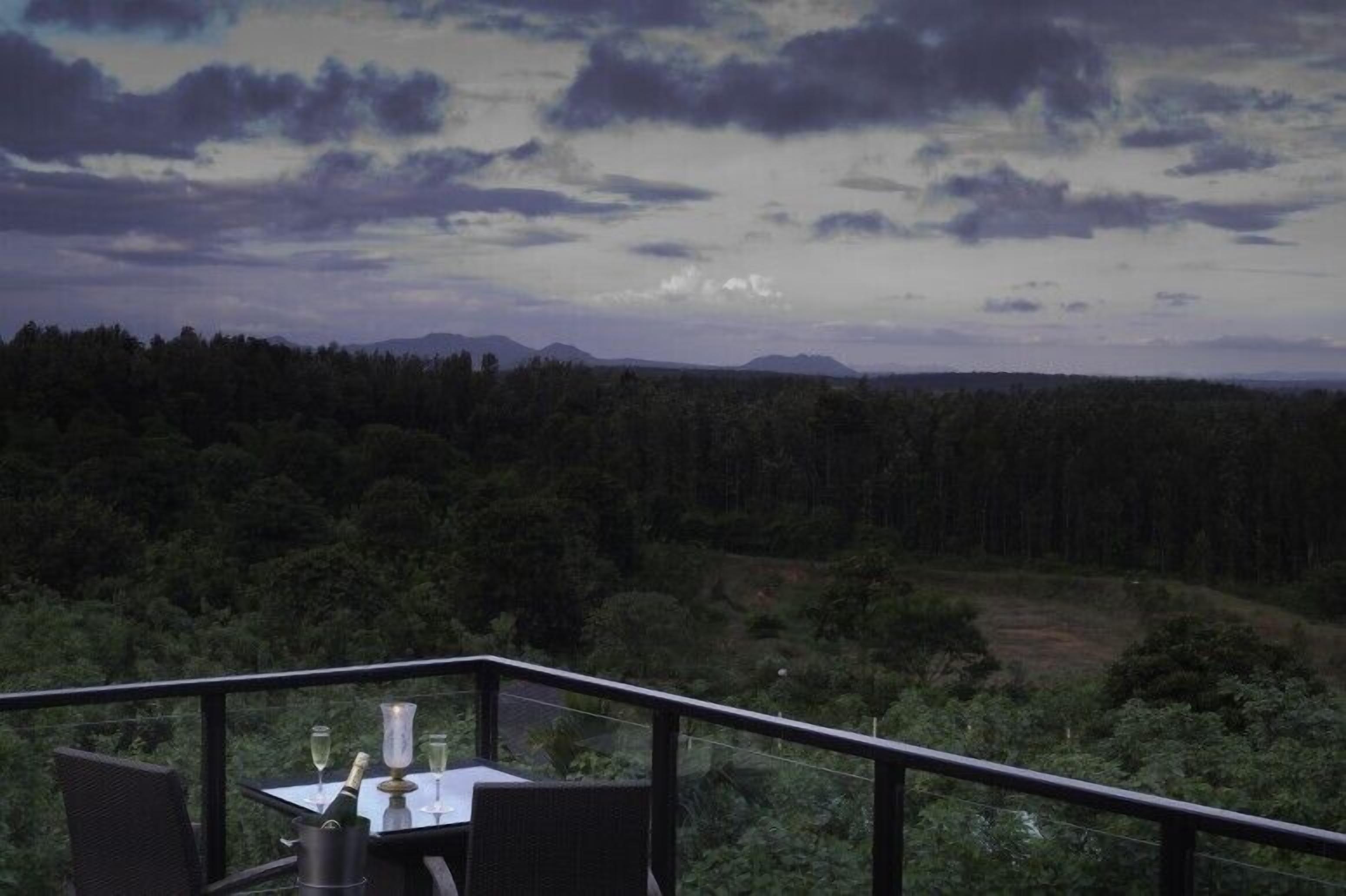 A balcony with a table and chairs, overlooking a forested landscape with mountains in the distance.