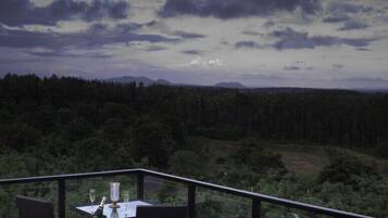 A balcony with a table and chairs, overlooking a forested landscape with mountains in the distance.