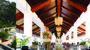 A spacious indoor area with a high ceiling and wooden beams, featuring a central walkway, potted plants, and a seating area with tables and chairs. - The Serai Chikmagalur (Chikkamagaluru)