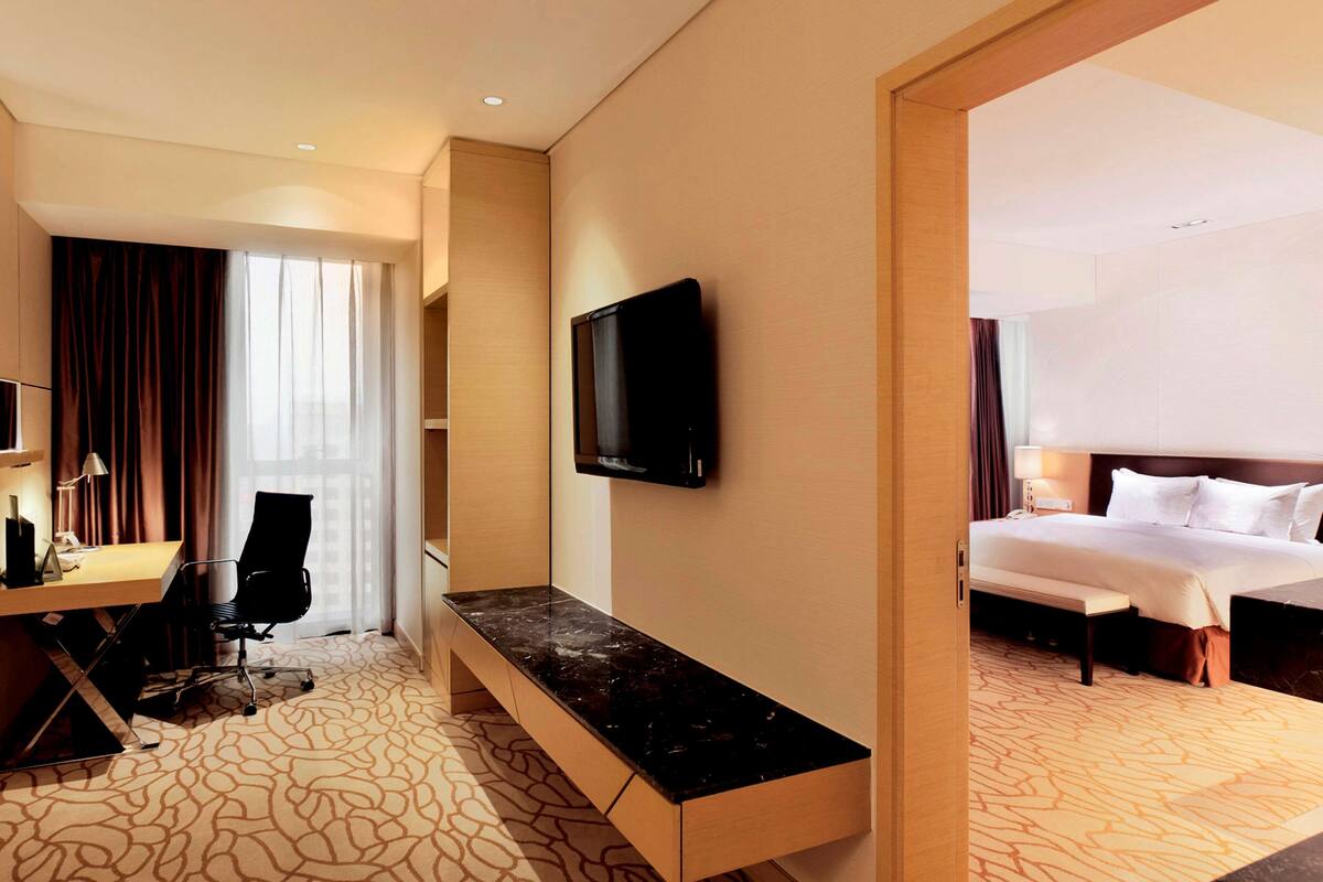 A hotel room with a bed, a desk with a chair, a TV mounted on the wall, and a bench.
