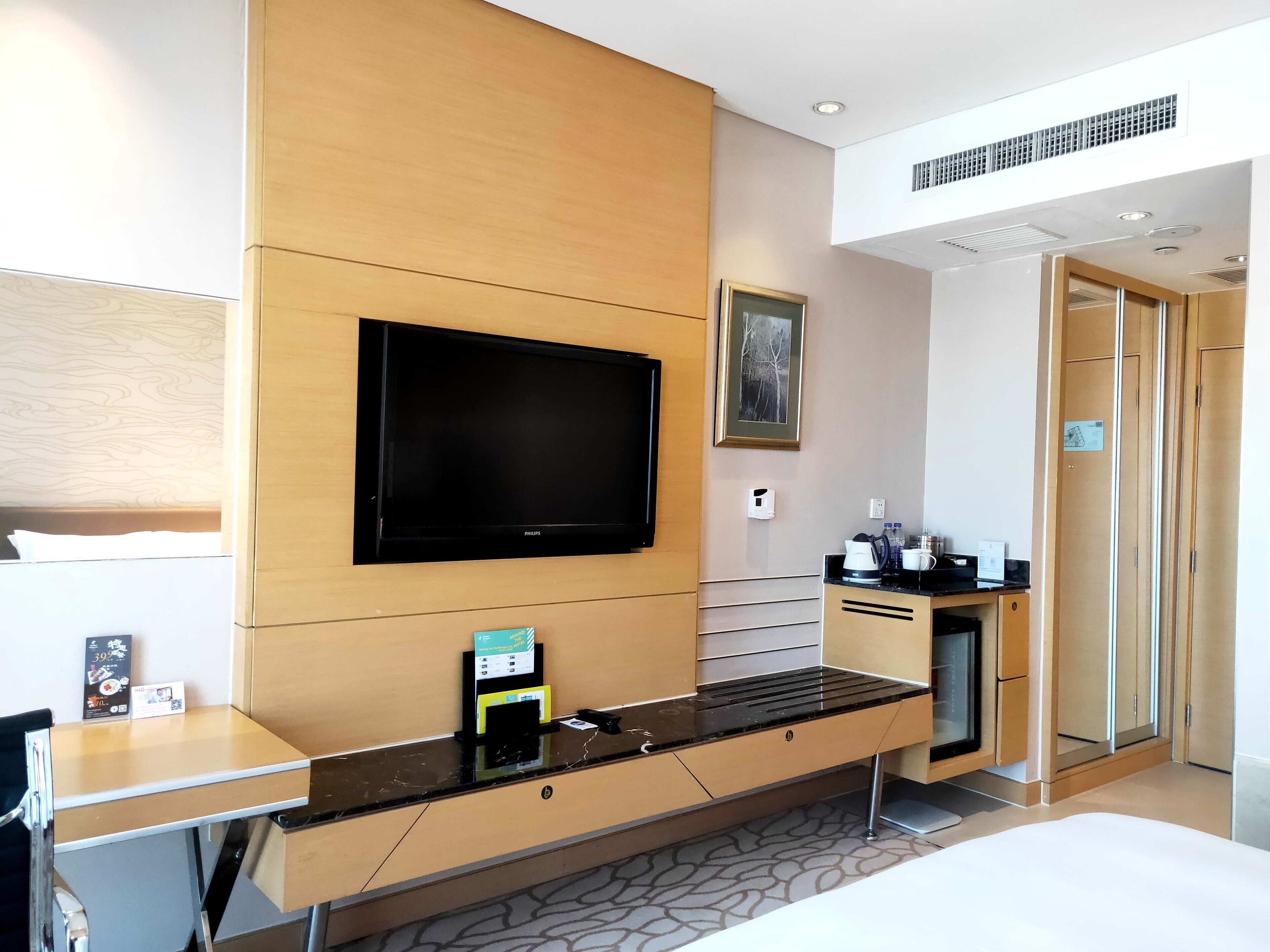 Habitación de hotel con televisor de pantalla plana montado en la pared, escritorio de madera con cafetera y cama con alfombra estampada.