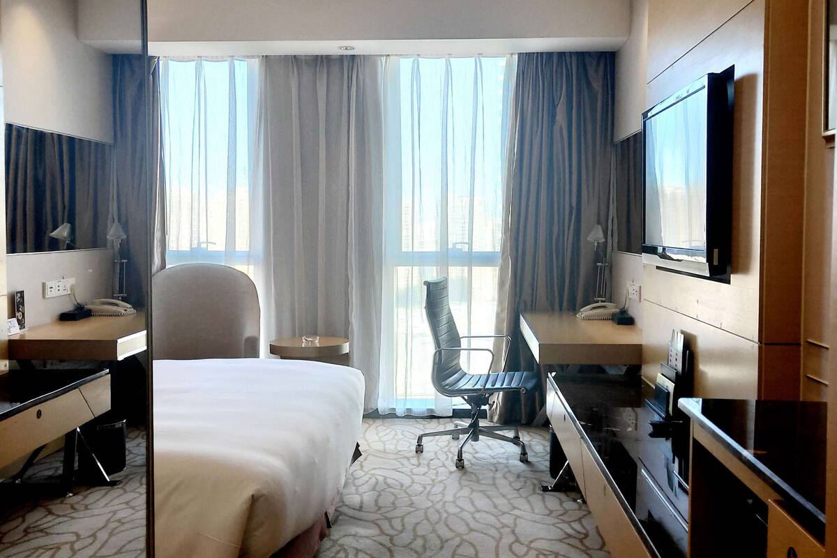 A hotel room with a bed, desk, chair, television, and large windows with curtains.