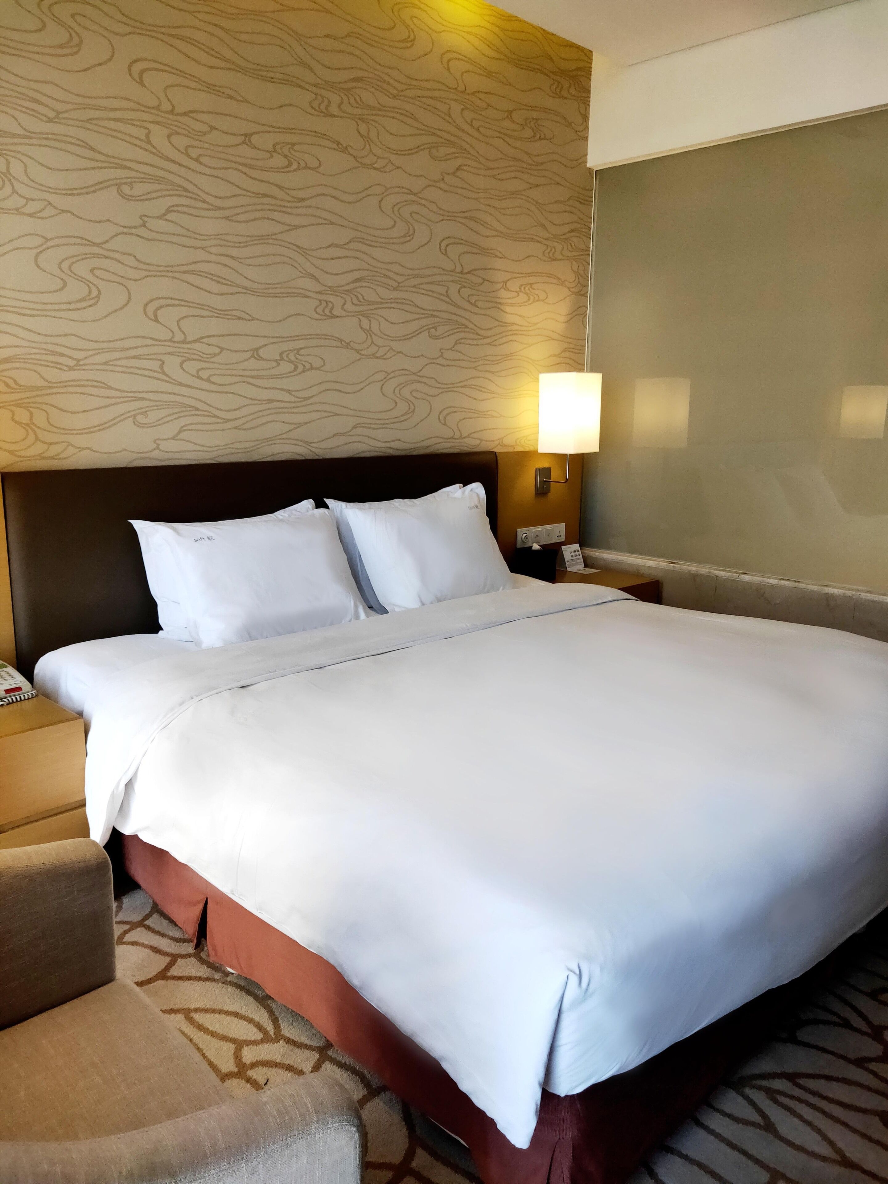 A hotel room with a large bed, a headboard with a wood grain pattern, a bedside lamp, and a small armchair.