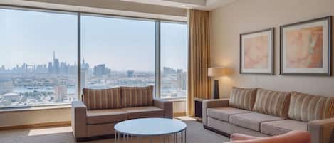 A spacious hotel room with a large window offering a city view, two sofas, a round coffee table, and a lamp.