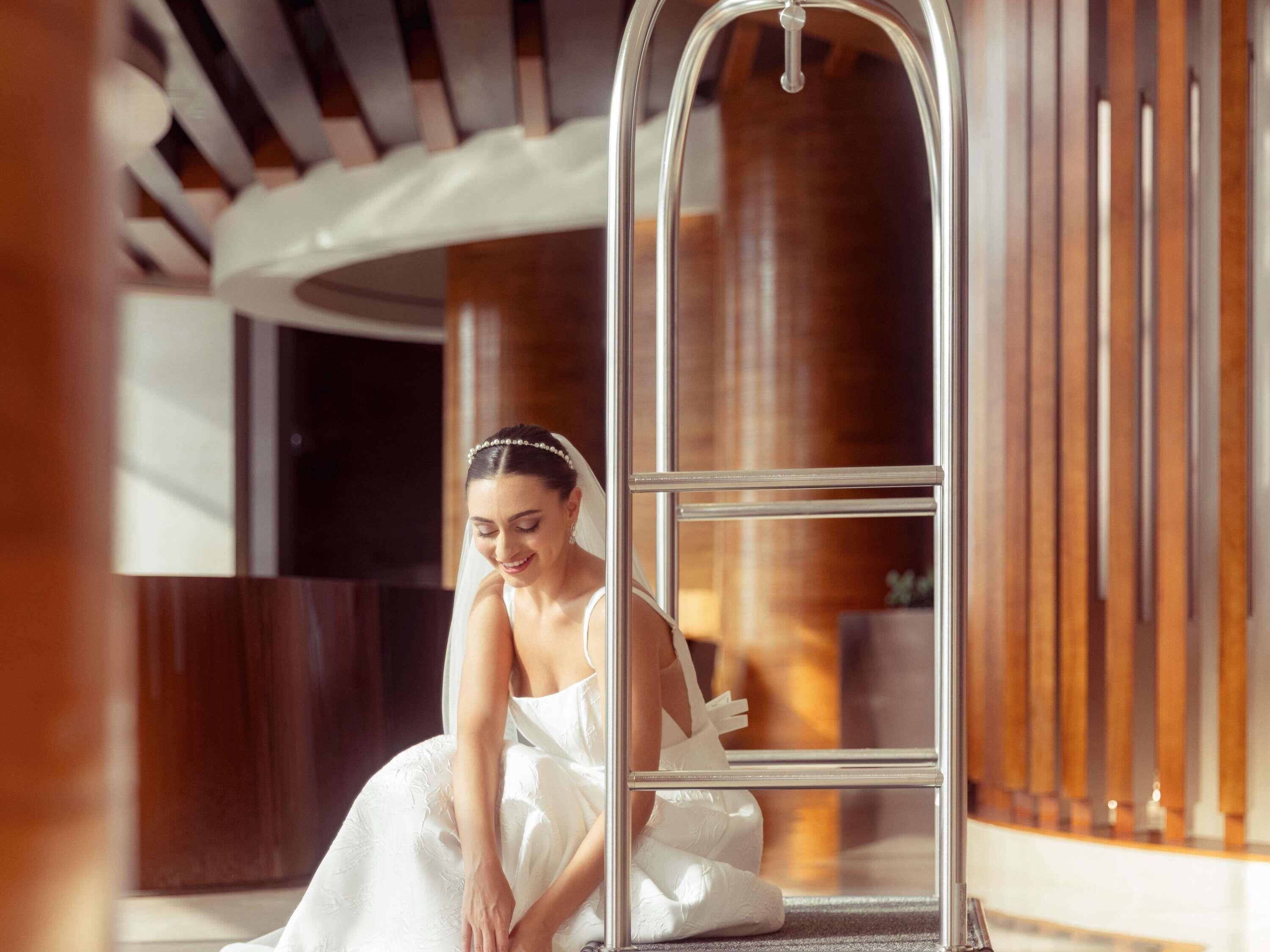 A person in a white wedding dress sitting on a metal bench in a modern indoor setting with wooden paneling and a glass door.