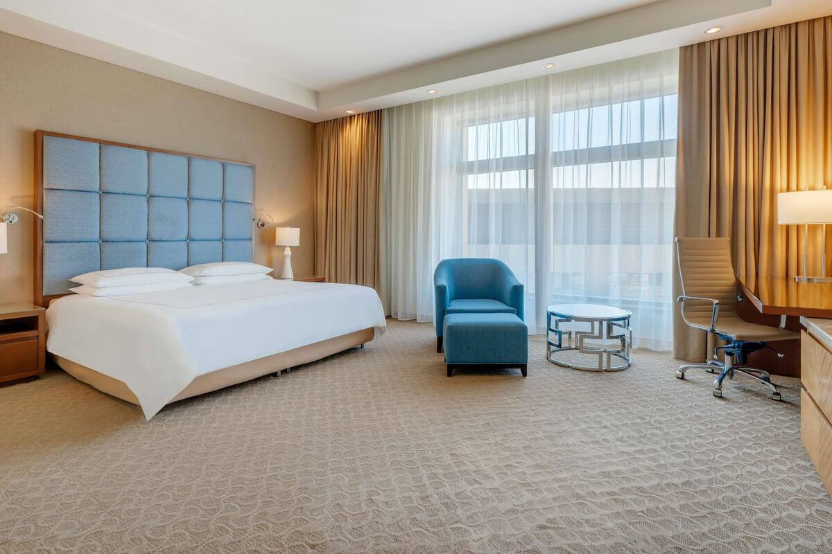 A hotel room with a large bed, a blue armchair, a small round table, and a desk with a lamp.