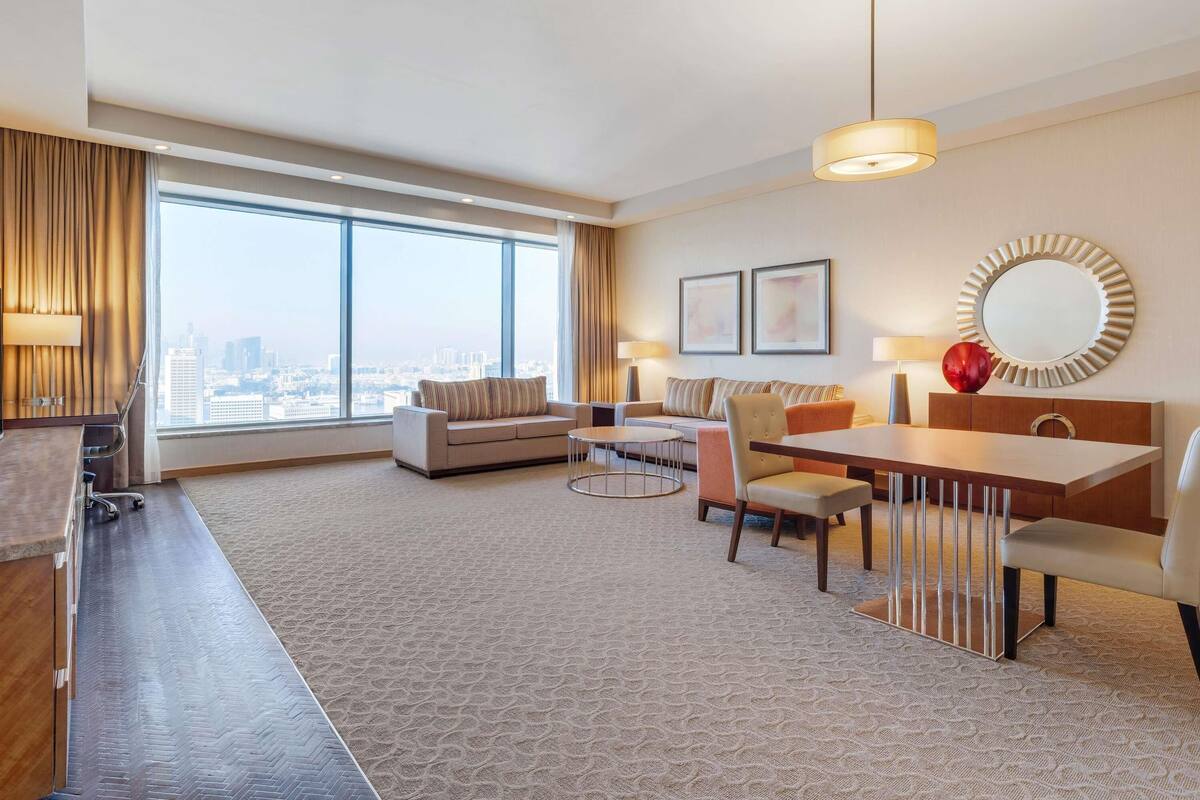 A hotel room with a large window, a sofa, a dining table, and a mirror.