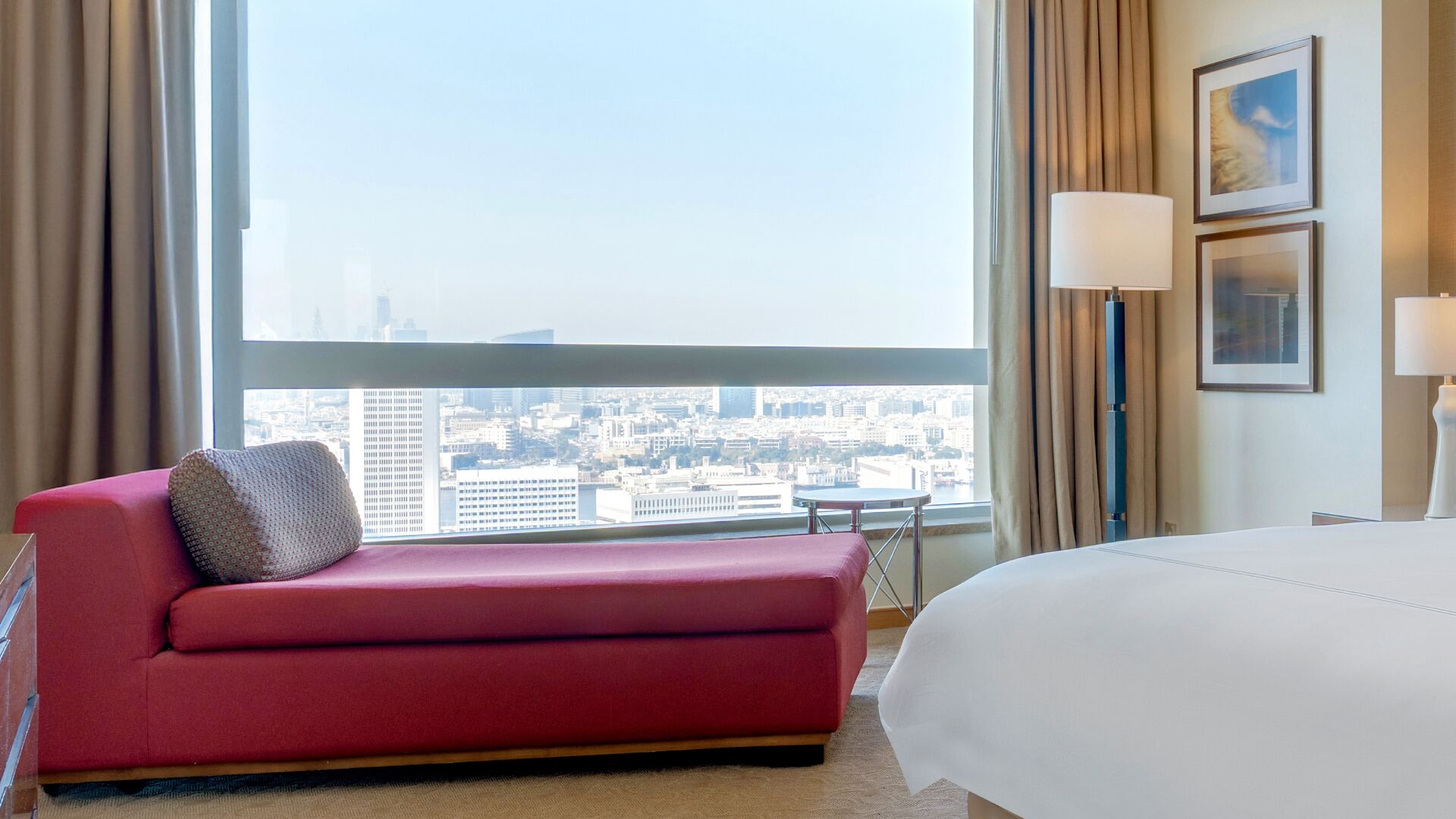 A hotel room with a large window, a red sofa, a bed, and two bedside lamps.