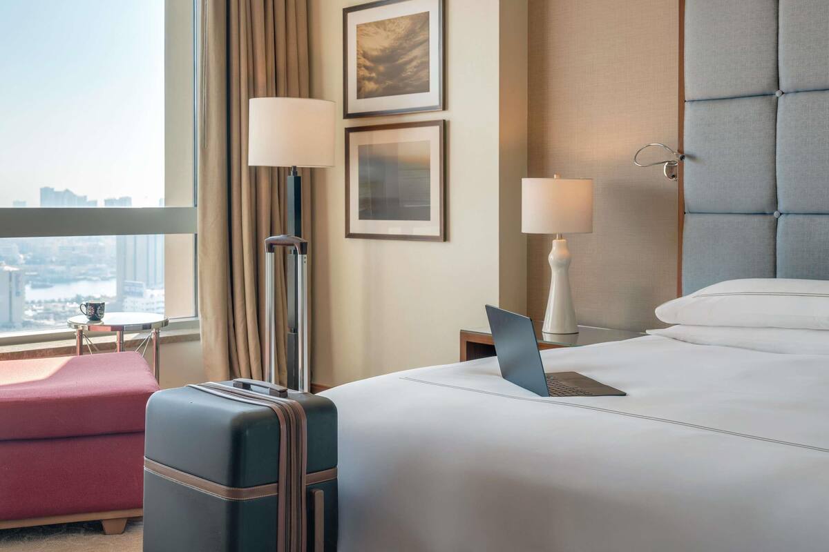 A hotel room with a bed, a desk with a laptop, a suitcase, a pink ottoman, and a view of the city.