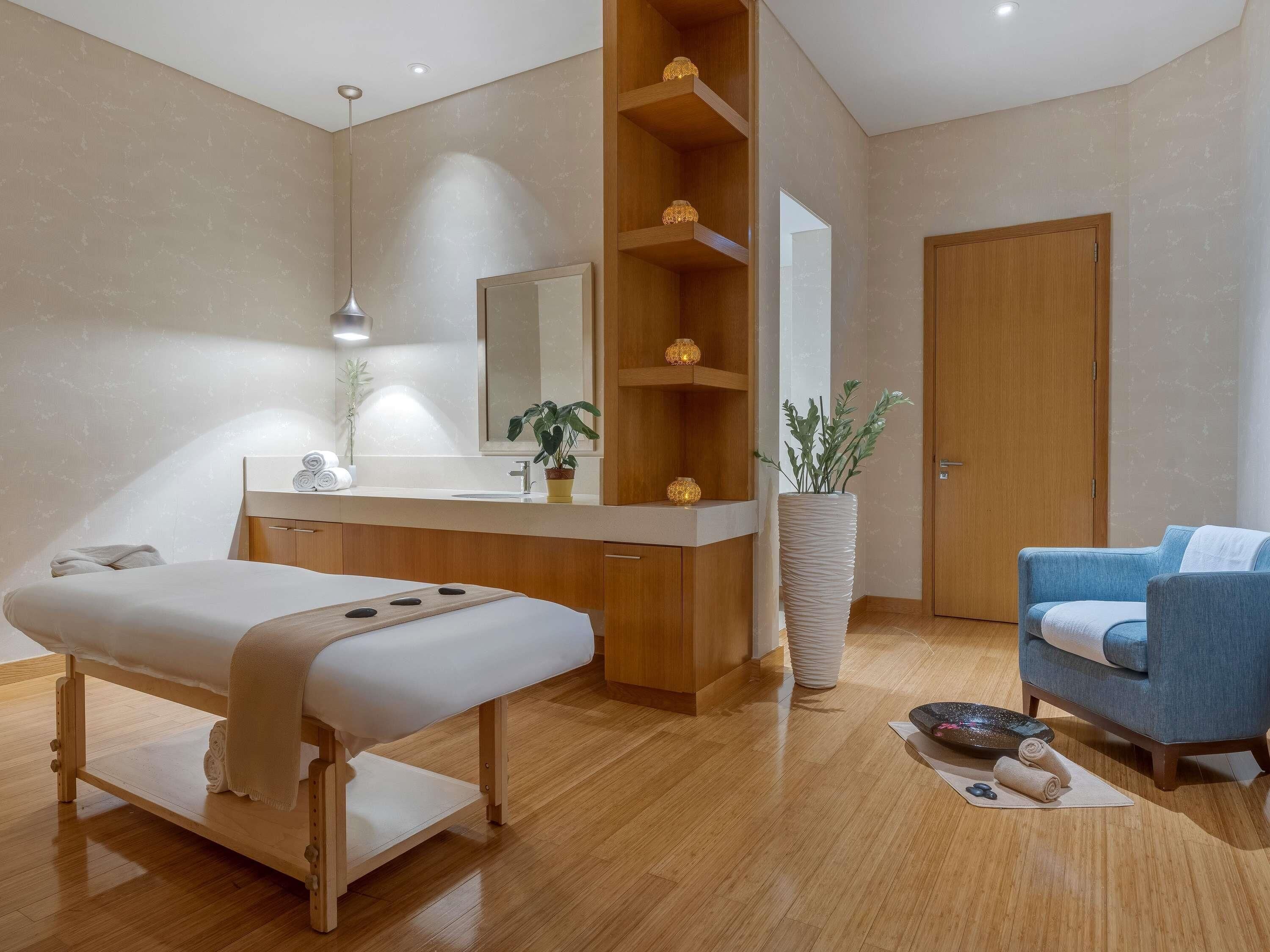 Un moderno spa con una camilla de masaje de madera, un mueble de madera, un espejo grande, una planta en maceta y una silla azul.