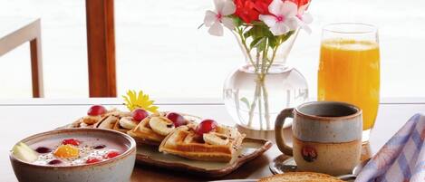 Desayuno con waffles, tostadas, tocino, un tazón de sopa, un vaso de jugo de naranja y un jarrón de flores.