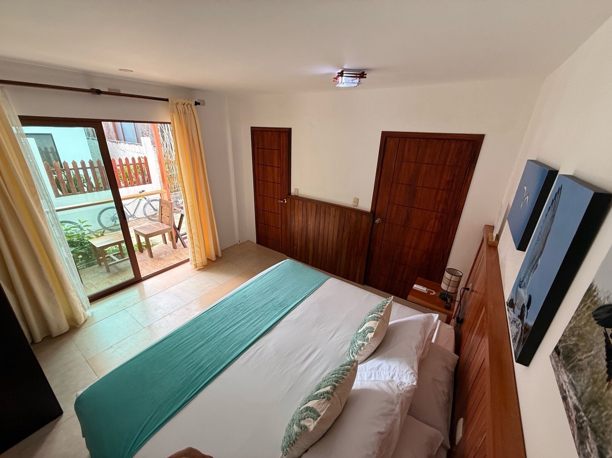 A bedroom with a bed, a wooden wardrobe, a sliding glass door leading to a balcony, and a small table with a lamp.