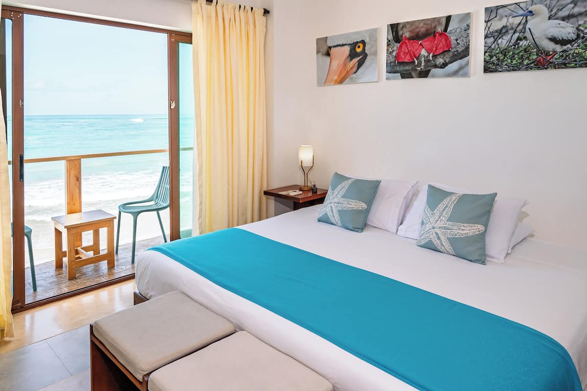 A hotel room with a bed, a view of the beach, a balcony with chairs, and artwork on the wall.