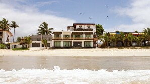 Un hotel frente a la playa con varios pisos, balcones y una playa de arena en primer plano.