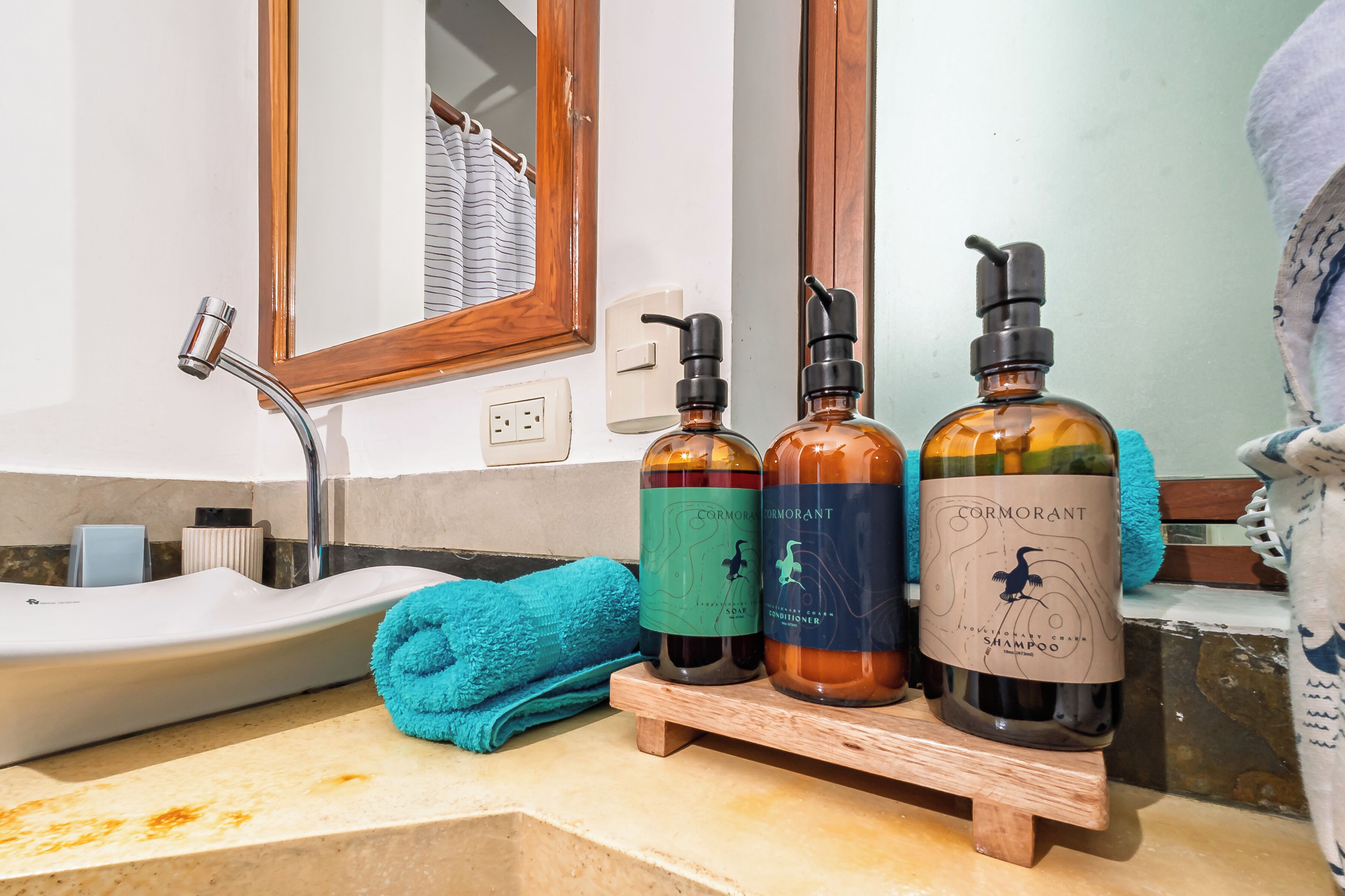 Une salle de bain avec un lavabo, un miroir et trois bouteilles de shampoing Cormorant, posées sur un présentoir en bois.
