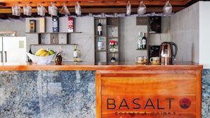 Mostrador de café y bebidas con diseño de madera, estantes con botellas y vasos, y un bol de frutas.