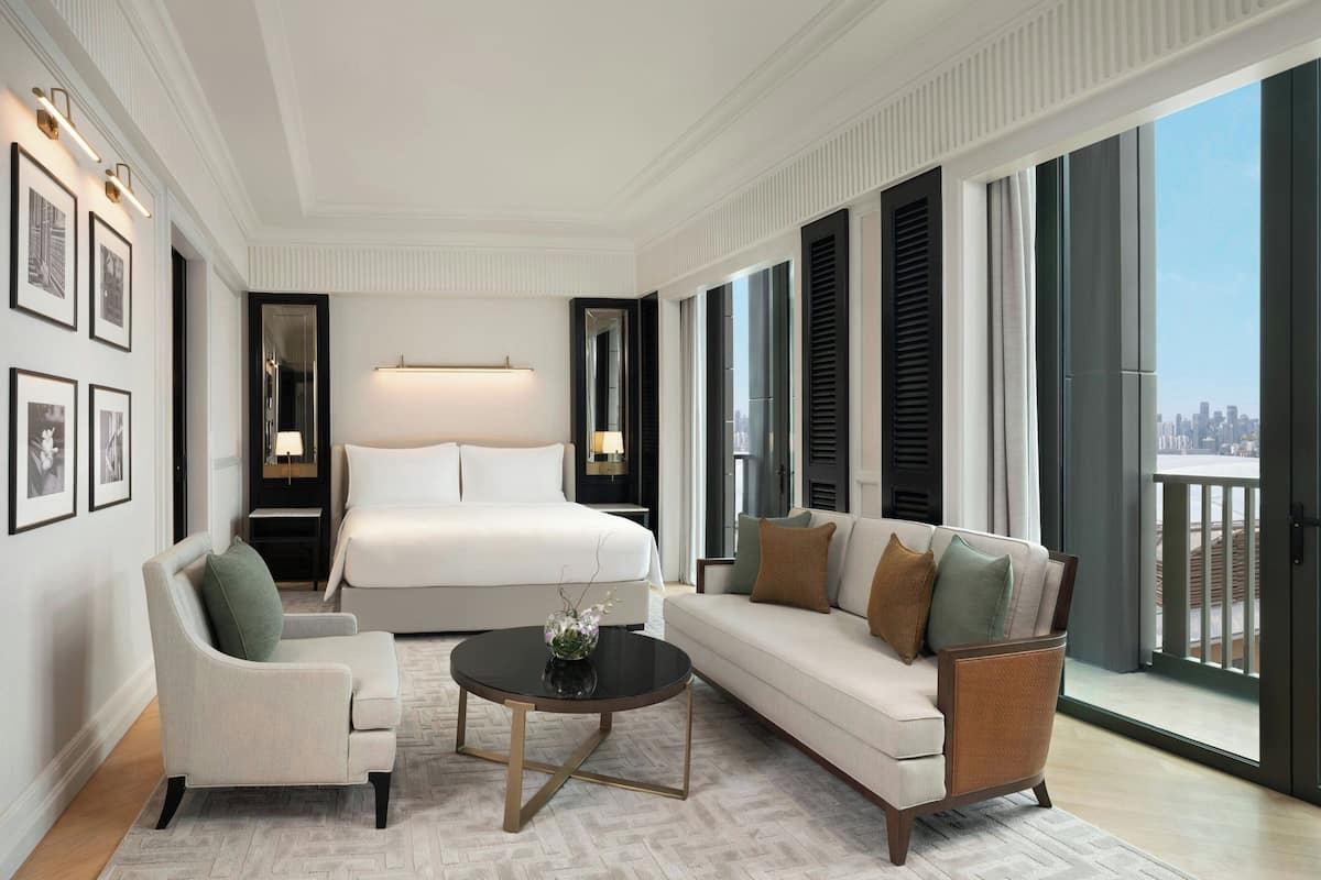 Een moderne hotelkamer met een groot bed, een bank, een stoel en een salontafel. Er zijn grote ramen met uitzicht op de stad.