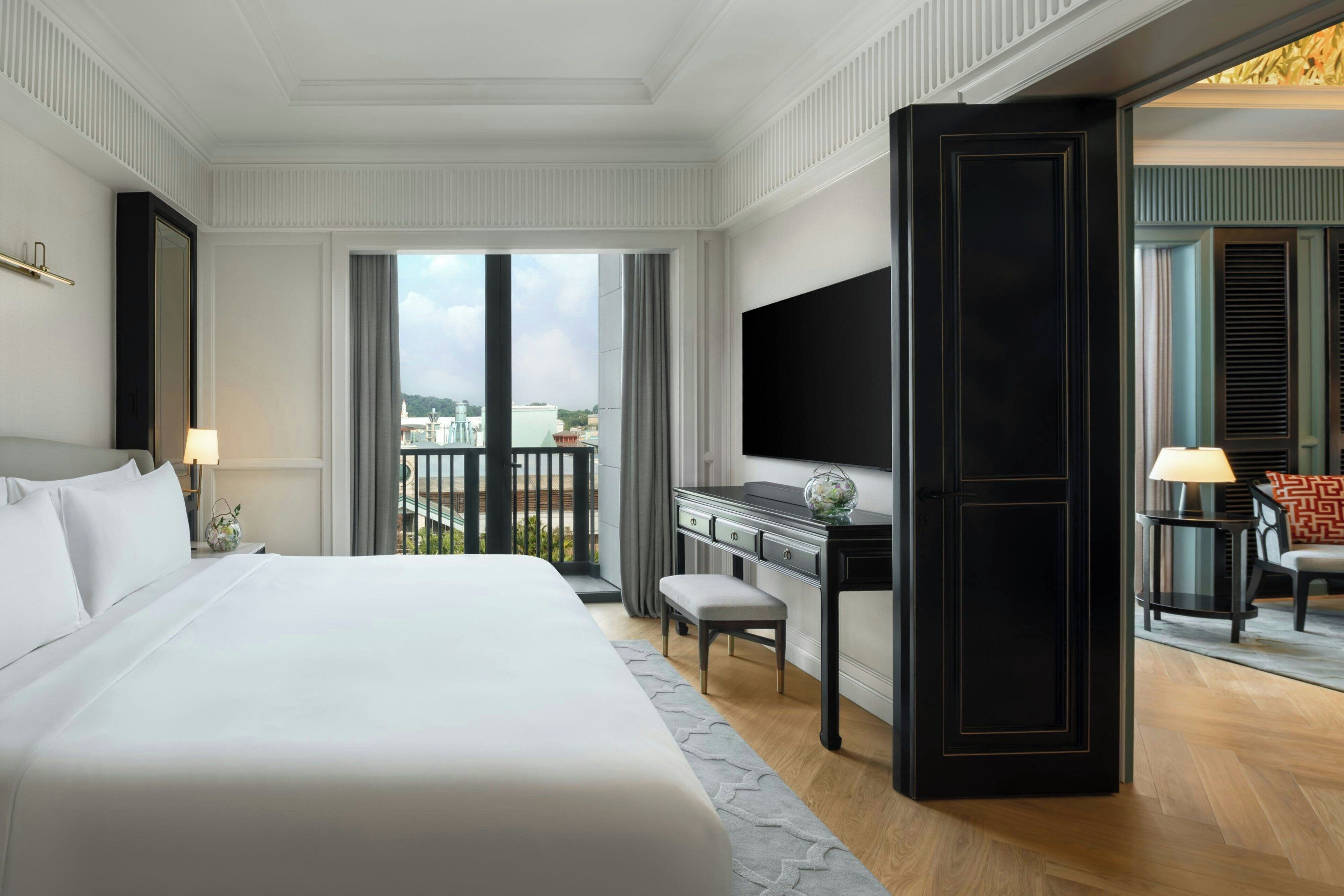 A hotel room with a large bed, a desk, a TV, and a balcony with a view of the city.