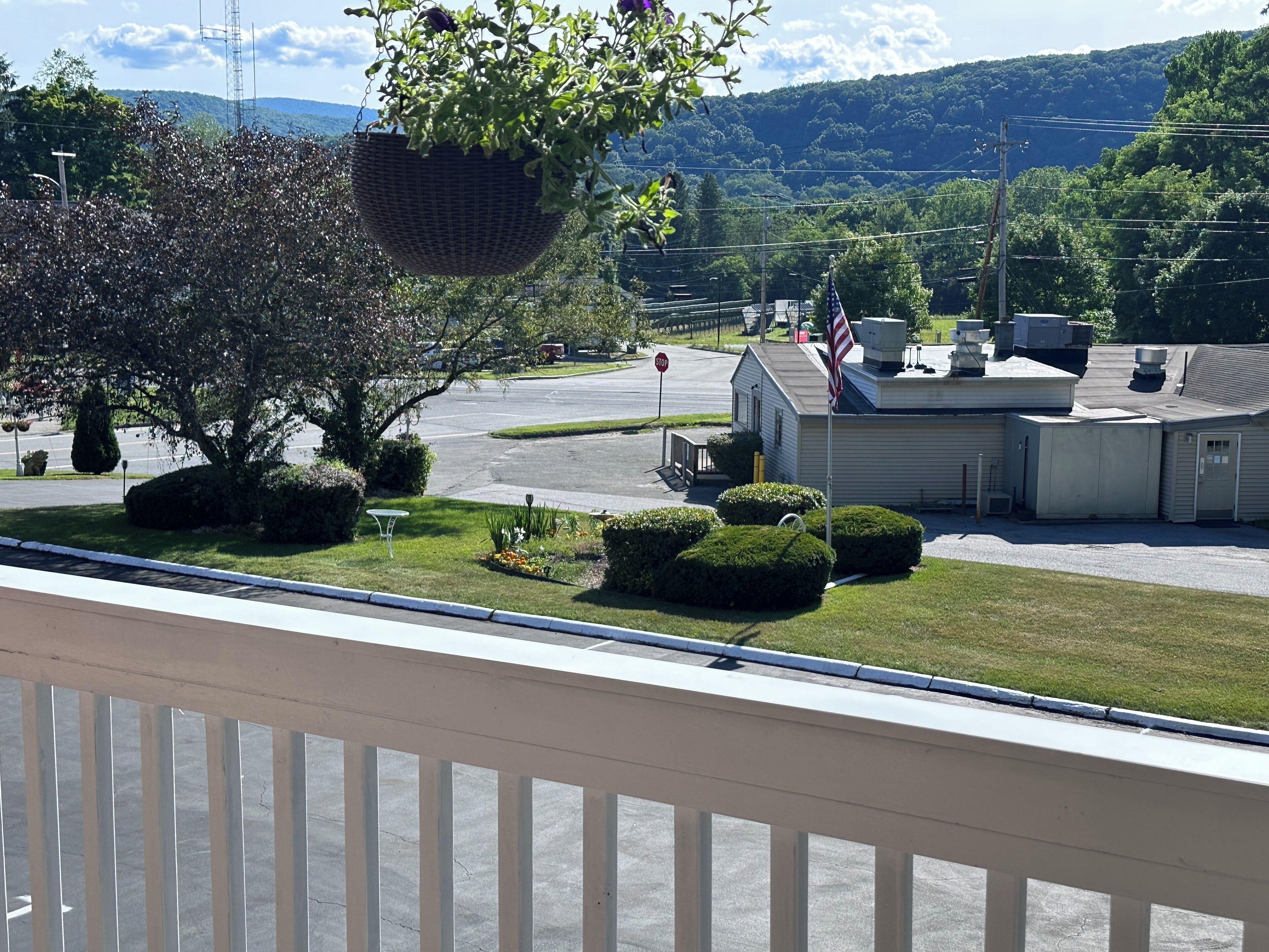 A view of a building with a parking lot, a stop sign, and a flagpole with an American flag. There are trees and shrubs in the foreground, and a mountain range in the background.