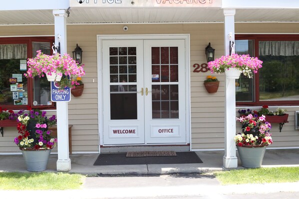 Un bâtiment doté d’une double porte sur laquelle on peut lire les mots « BIENVENUE » et « BUREAU », d’un panneau indiquant « PARKAGE RÉSERVÉ AUX AUTORISÉS » et de jardinières de chaque côté de l’entrée.