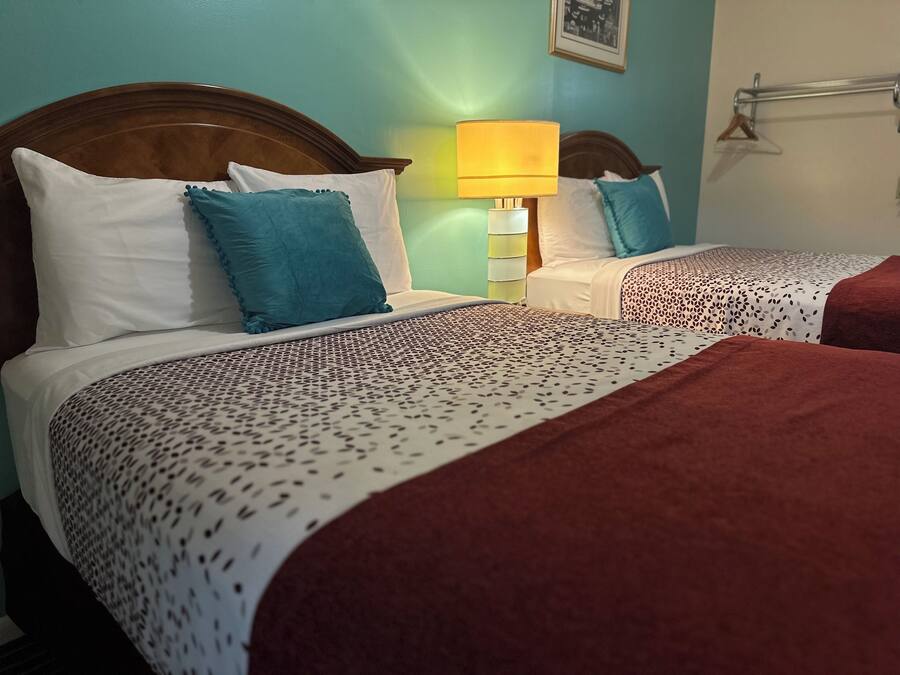 Two beds with patterned bedspreads, a nightstand with a lamp, and a wall-mounted towel rack.