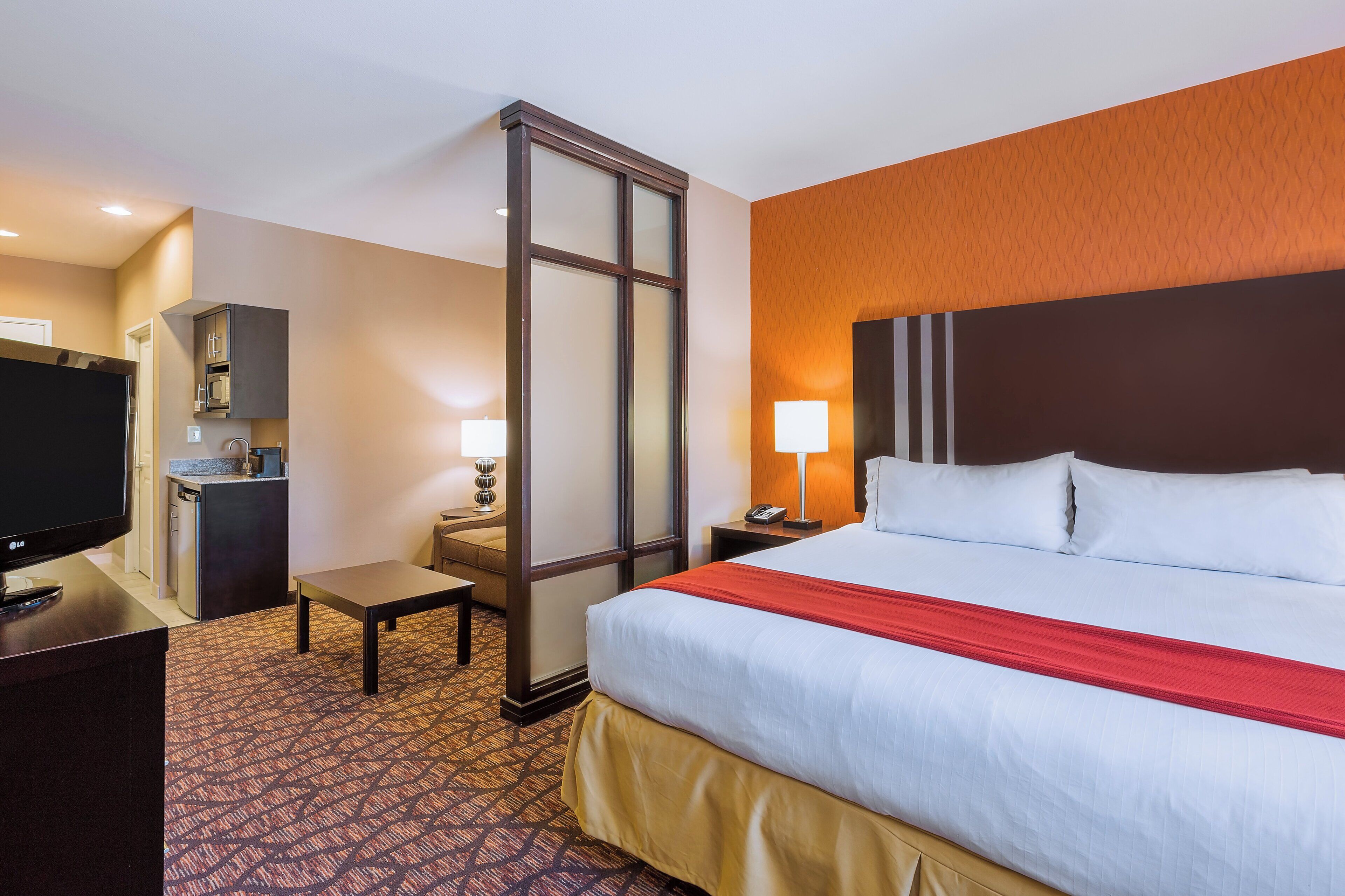 A hotel room with a bed, a desk with a television, a small table, a nightstand with a lamp, and a bathroom door.
