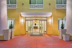 Entrada de hotel con puerta de vidrio, camino de ladrillo y plantas en macetas a ambos lados.