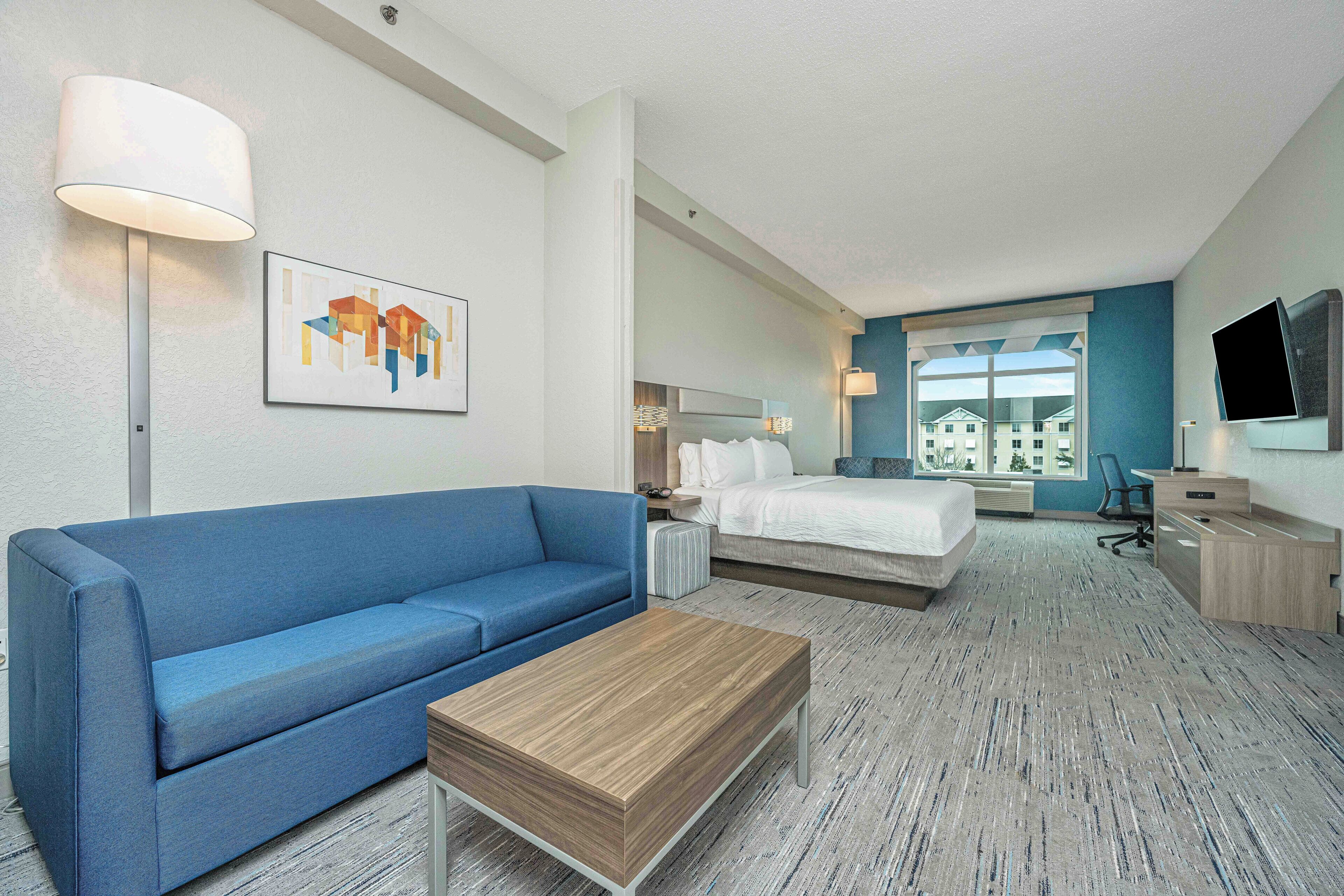Habitació d'hotel amb un sofà blau, una taula de centre de fusta, un llit amb roba de llit blanca, un escriptori amb un televisor de pantalla plana i una finestra gran amb vista a edificis.