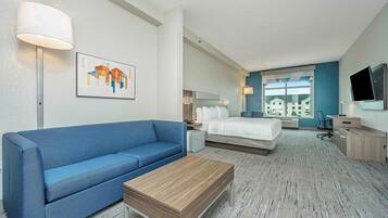 Quarto de hotel com sofá azul, mesa de centro de madeira, cama com roupa de cama branca, escrivaninha com televisão de tela plana e uma janela grande com vista para prédios.