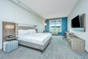 A hotel room with a large bed, a desk, a chair, a TV, and a window with a view of buildings. - Holiday Inn Express & Suites Charleston Arpt-Conv Ctr Area (North Charleston)