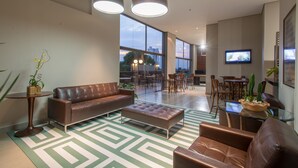 A modern living room with a brown leather sofa, a glass coffee table, and a patterned rug. There is a television mounted on the wall and a plant on the coffee table. - Hotel Gran Odara (Cuiabá)