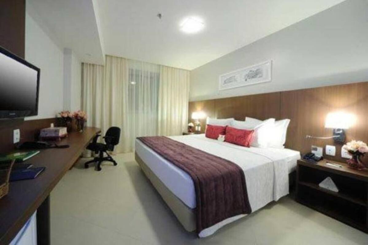 A hotel room with a bed, desk, chair, TV, bedside lamps, and a framed picture on the wall.