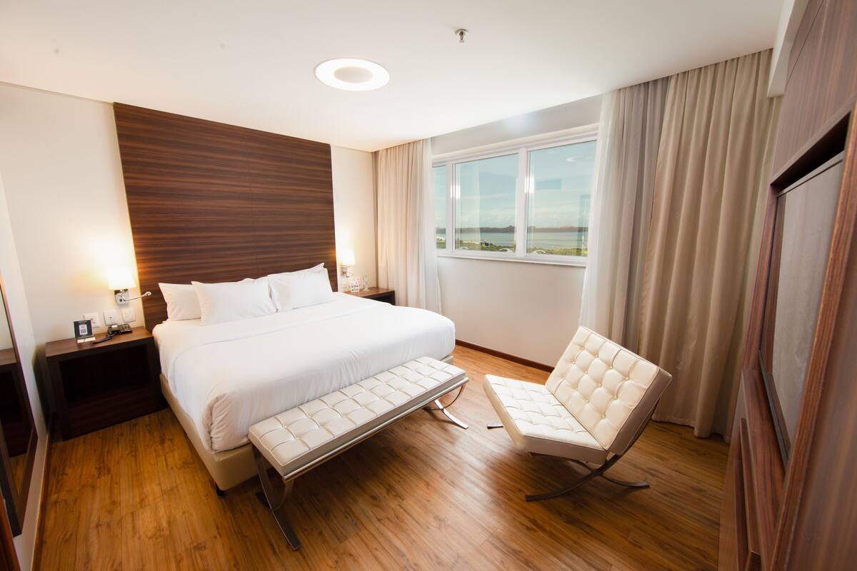 A modern hotel room with a large bed, a bedside table, a chair, a window with a view, and a wooden floor.